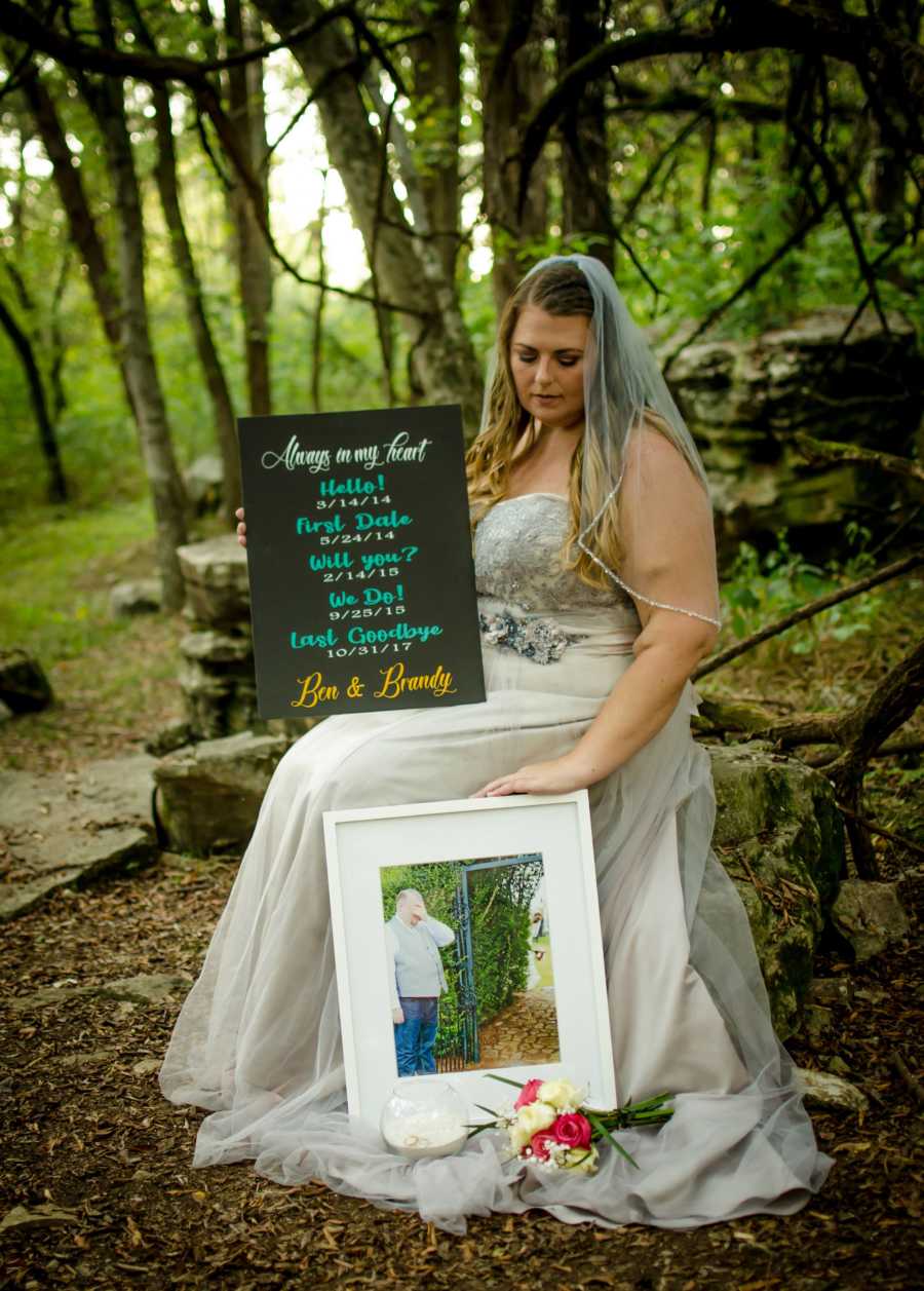 Woman in wedding dress sits in woods holding sign for deceased husband beside picture of him