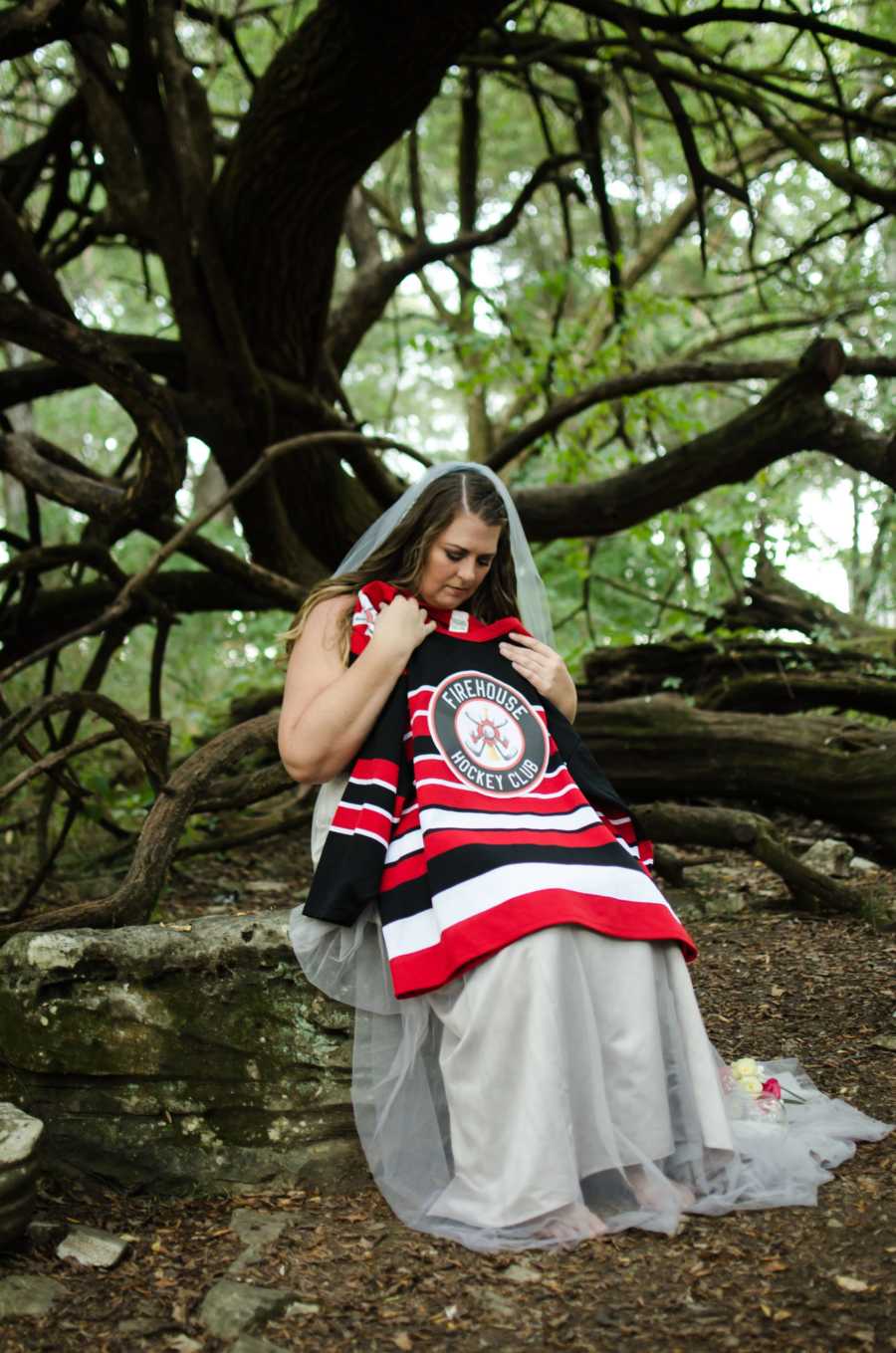 Woman sits on tree trunk in forest wearing wedding dress holding up late husband's hockey jersey