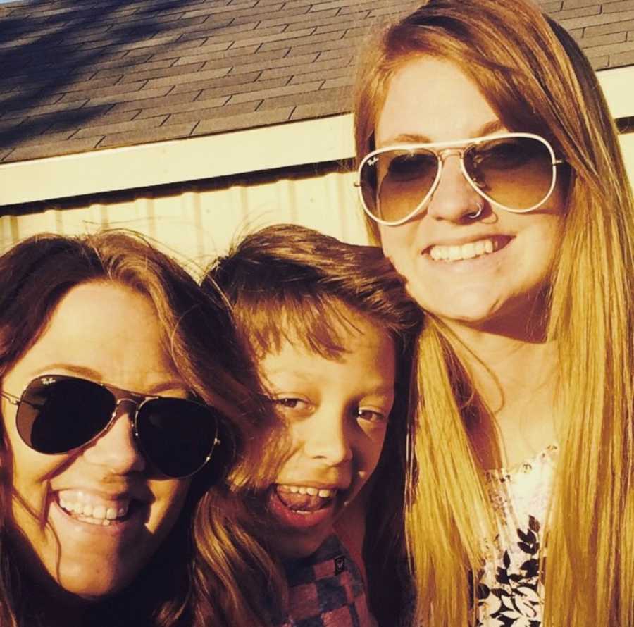 Young boy smiles outside with mom and step mom who are both wearing sunglasses