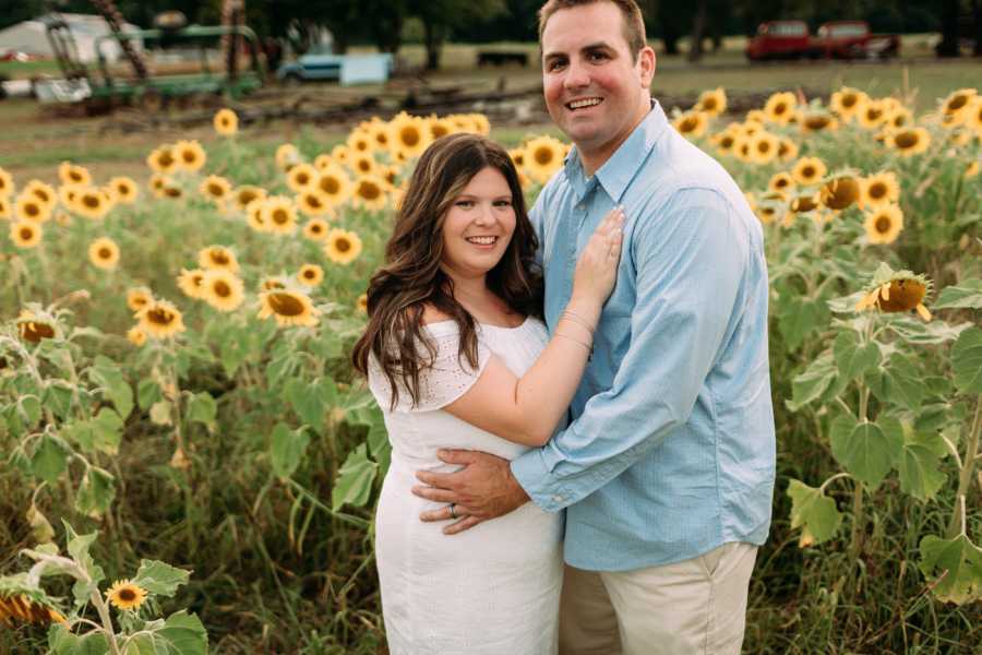 Pregnant woman stands in arms of husband in sunflower field