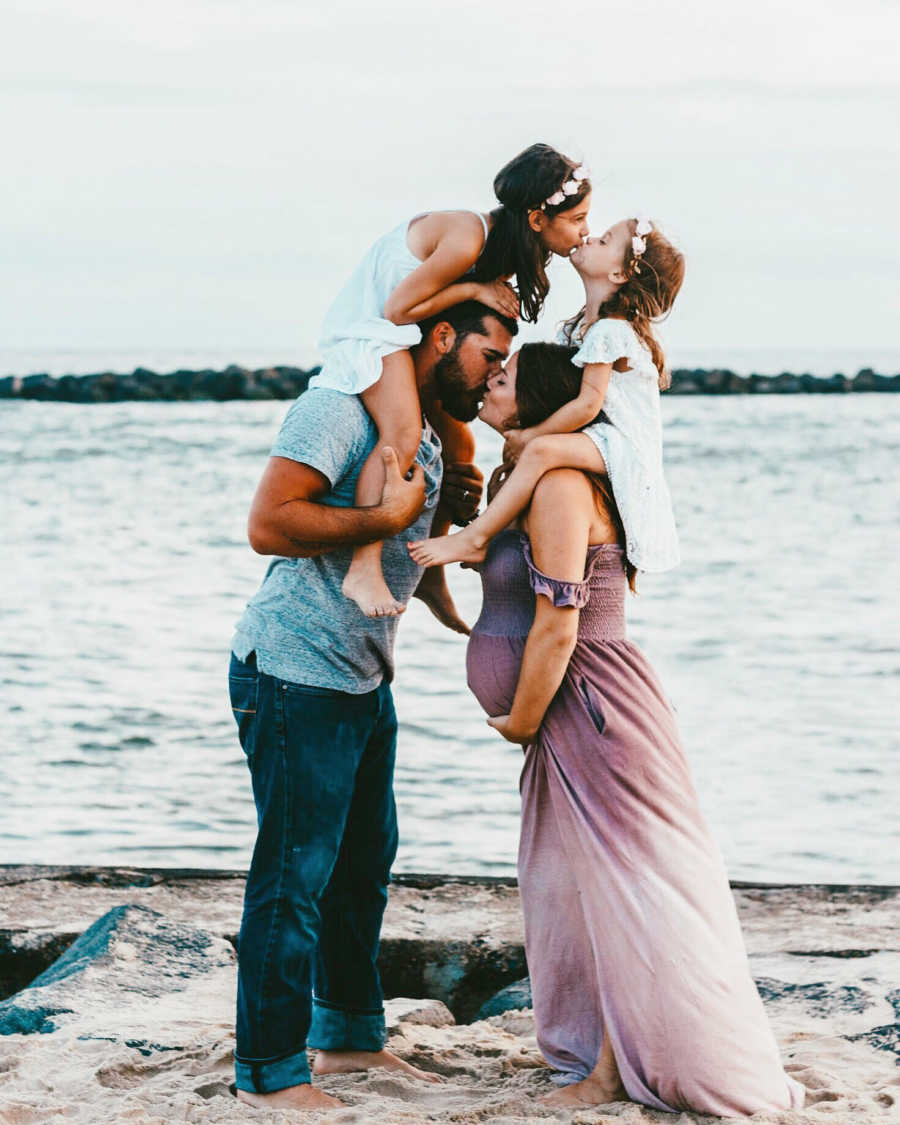 Husband and pregnant wife kiss on beach with their two daughters sitting on their shoulders
