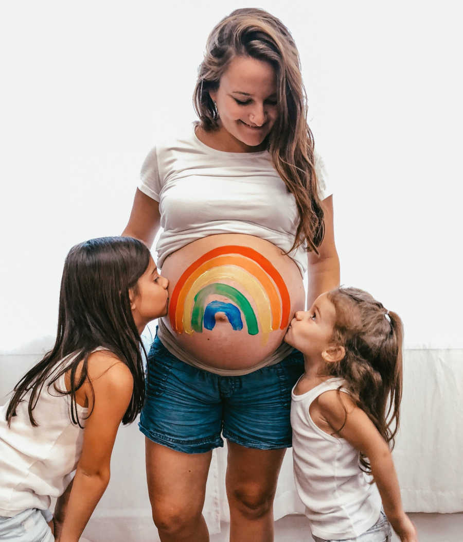 Pregnant mother with rainbow painted on her stomach stands while her two daughters kiss her stomach