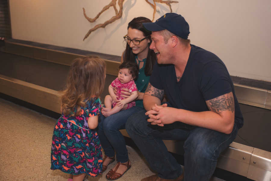 Husband and wife sit on luggage carousel with adopted baby in wife's lap and their older daughter standing beside them