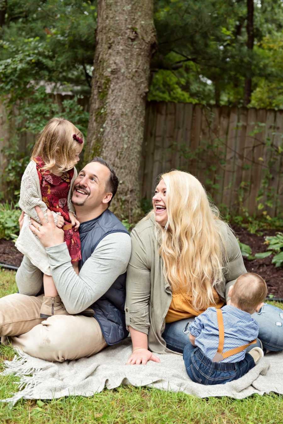 Mother and father sit on blanket outside with their biological daughter and adopted son