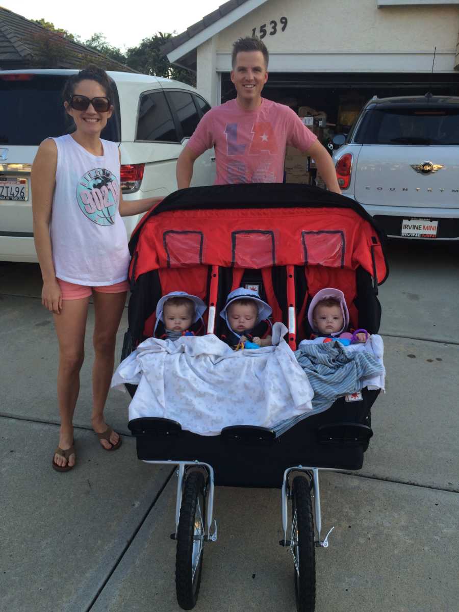 Husband stands beside wife in driveway holding stroller where his triplets sit