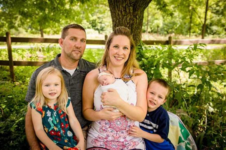 Husband and wife with newborn swaddled to her chest sit outside on bench with their young son and daughter