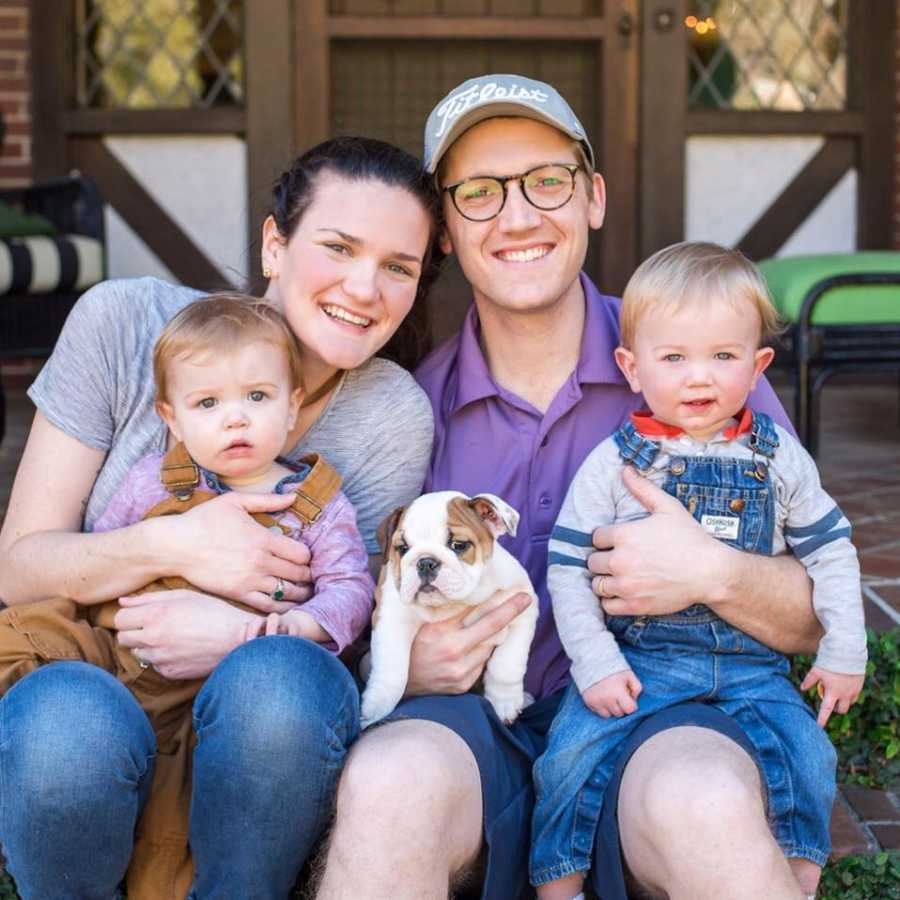 Husband and wife sit outside their front door with son and daughter in their lap and puppy between them