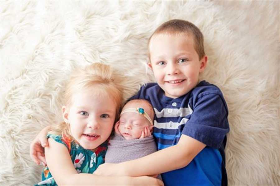 Older brother and sister lay on white carpet holding preemie sibling 