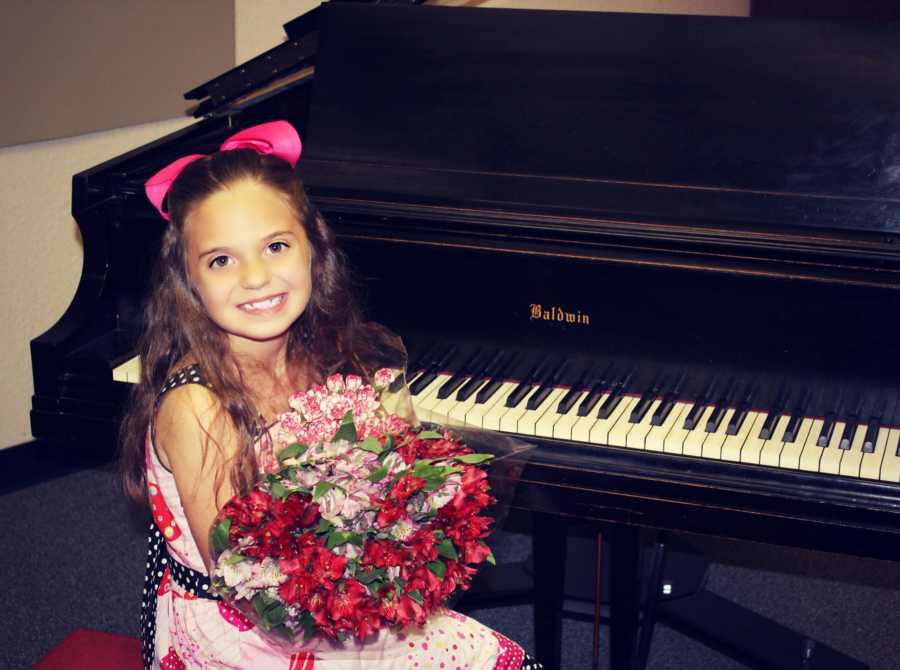 Little girl who has had a lot of medical issues sits smiling at piano holding bouquet of flowers