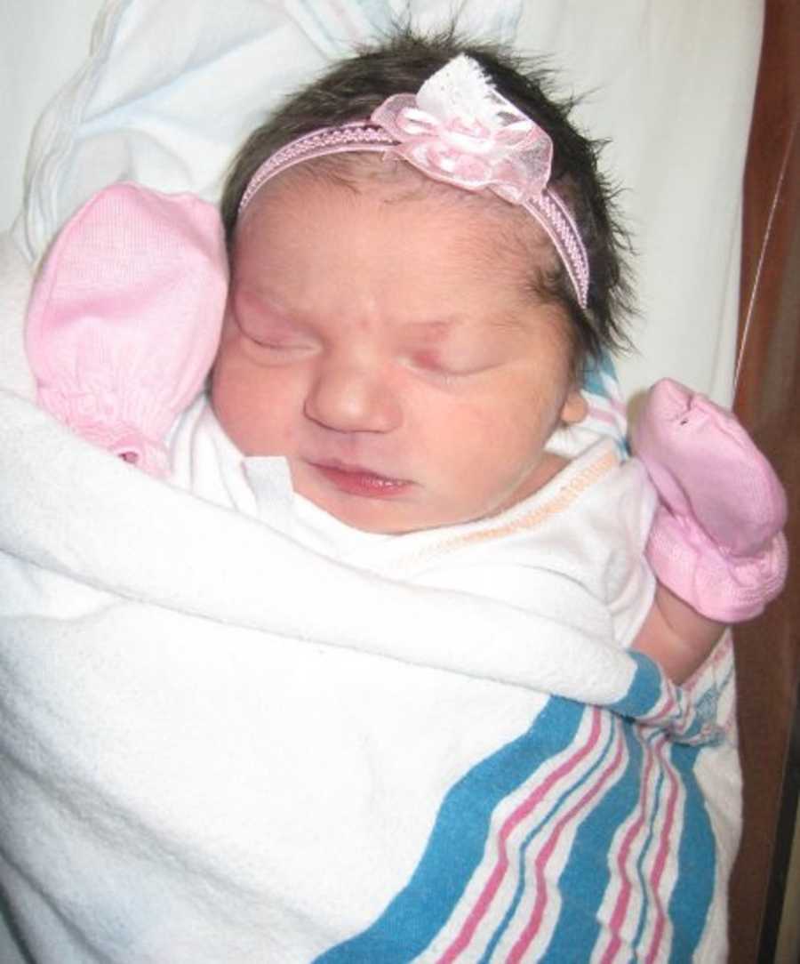 Newborn baby lays asleep swaddled in blanket wearing pink mittens and pink headband with bow