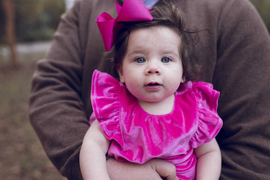 Adopted baby held in arms of her father in pink dress with big pink bow in her hair