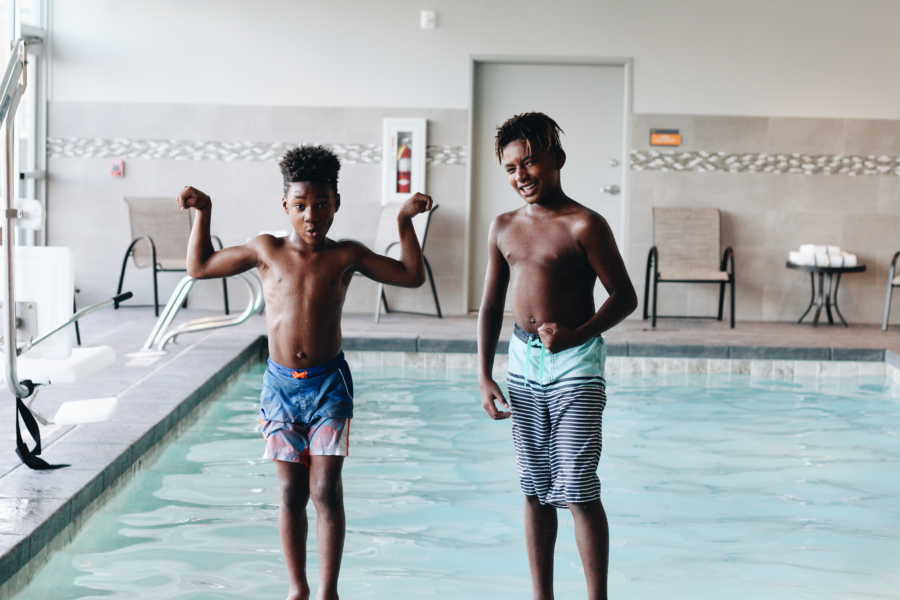 Young boy who was adopted at birth stands beside biological brother as they flex their muscles by indoor pool