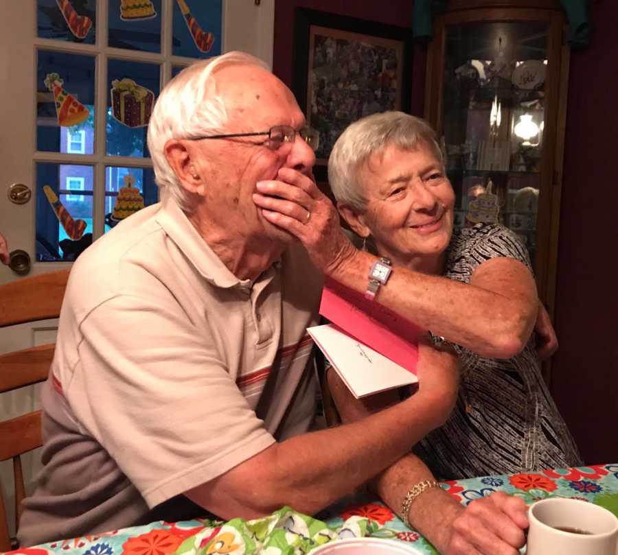 Woman with dementia sits at table beside husband in home holding hand over his mouth