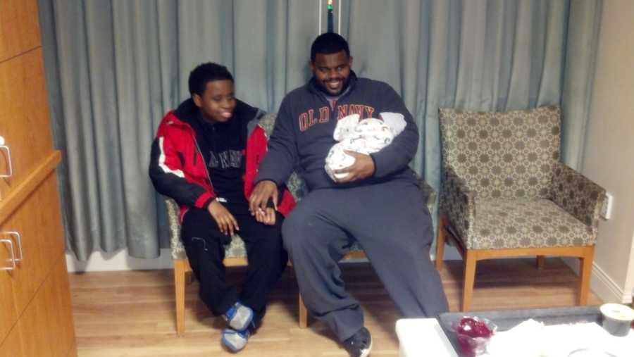 Father sits in hospital room holding newborn baby beside teen son