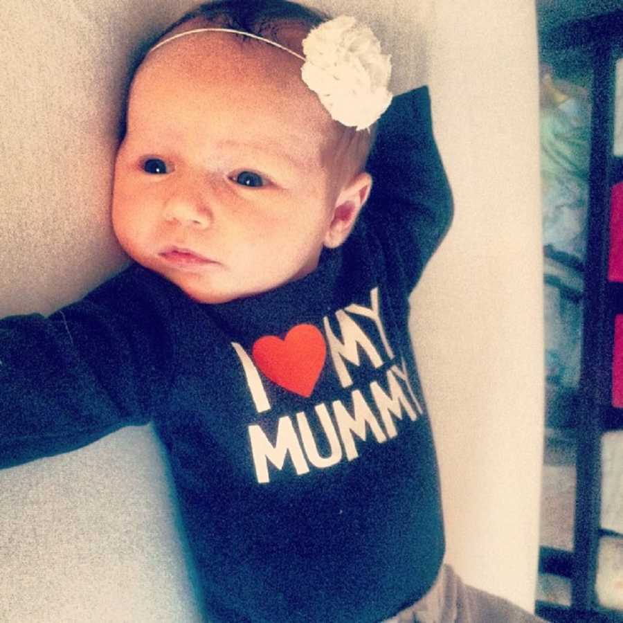 Baby girl lays on her back wearing shirt that says, "I heart my mummy" and white flower headband