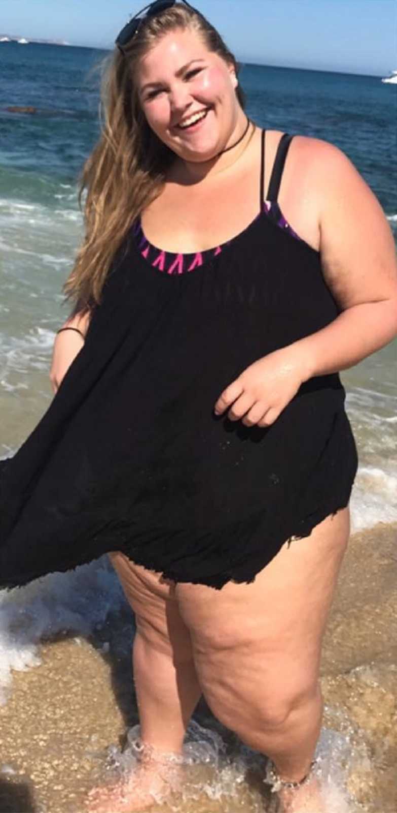 Teen who was bullied for her weight stands smiling in shallow water of ocean in coverup