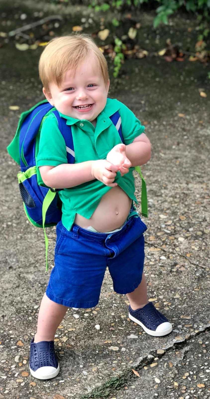 Toddler boy stands smiling with backpack on