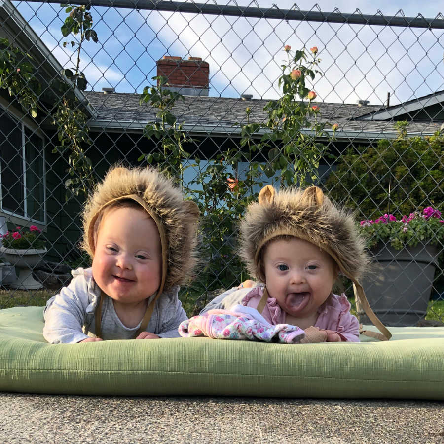 Twin girls with down syndrome lay outside on green pillow with lion mane hats on