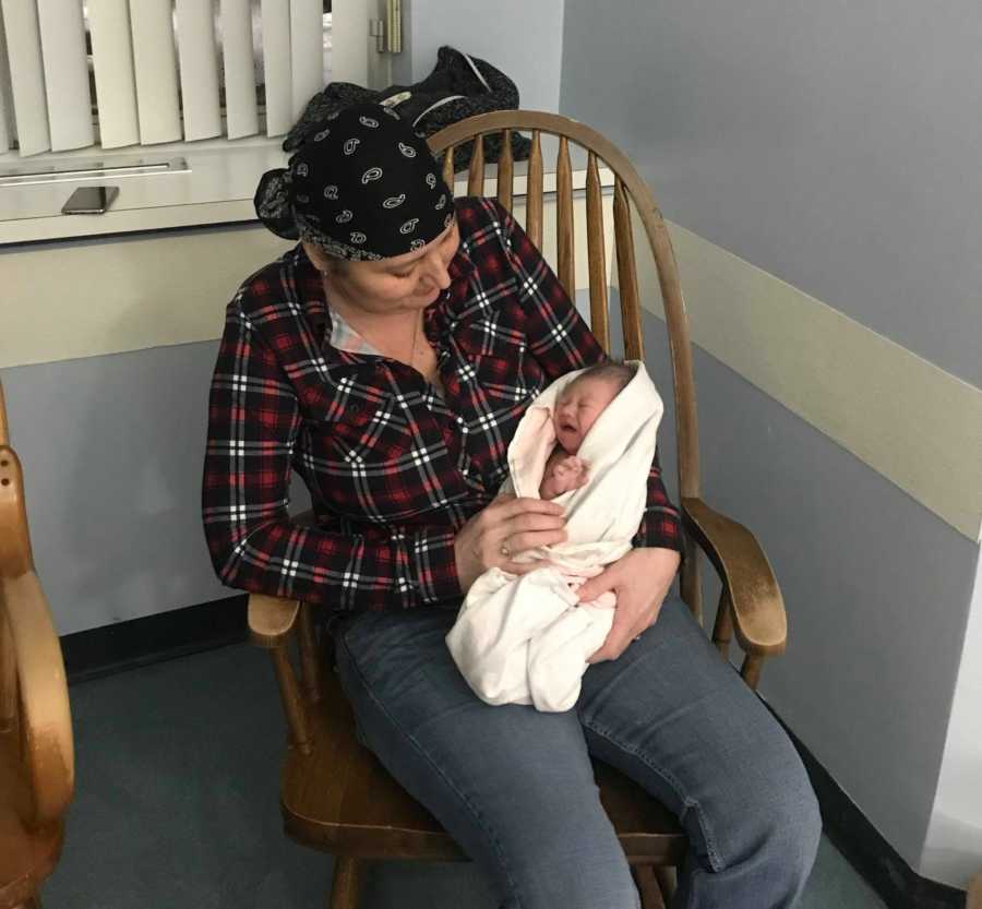 Woman with cancer sits in hospital chair holding newborn grandchild