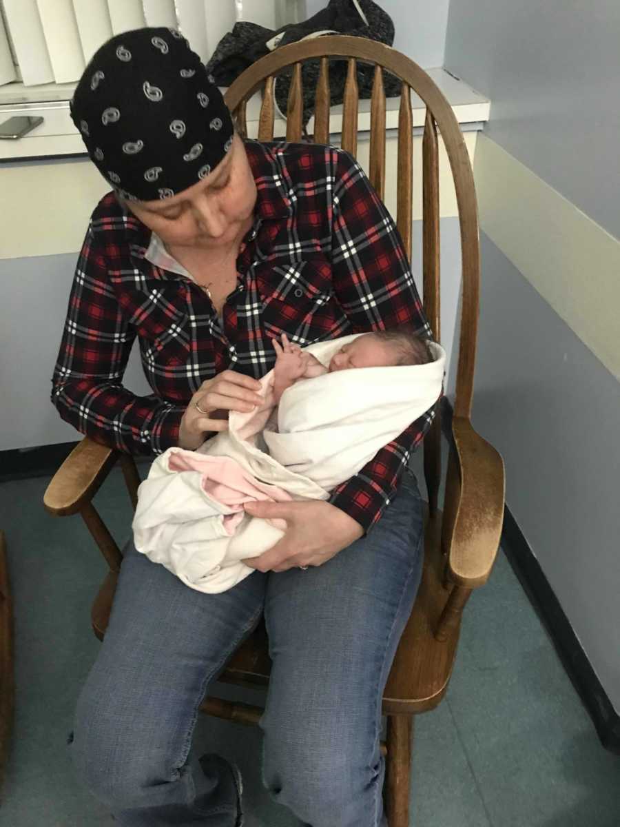 Grandmother with cancer sits in chair in hospital room holding newborn