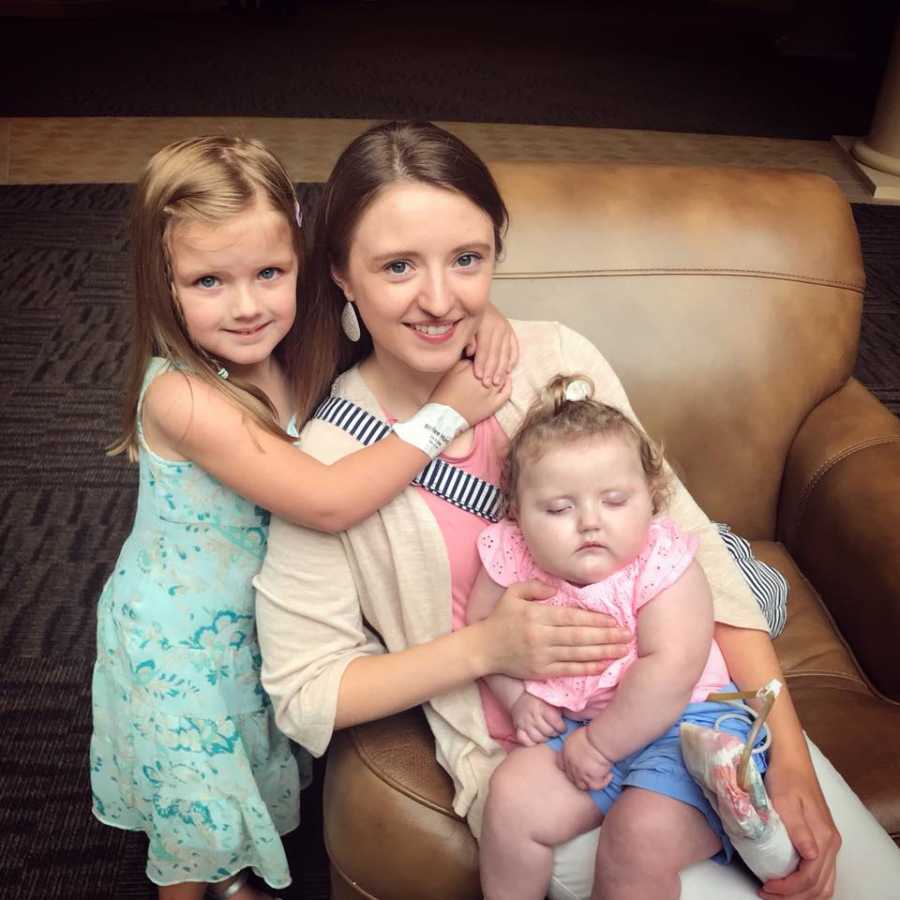 Mother smiles in chair with one daughter hugging her and other daughter with blindness, Cerebral Palsy, and Gastroparesis sits in her lap
