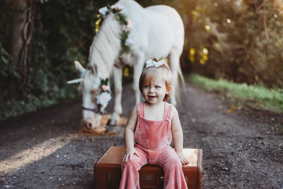 Toddler who was conceived through egg donor sits on wooden box in velvet onesie with horse behind him