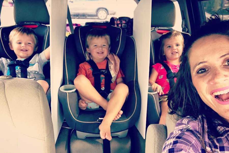 Mother smiles in selfie in her car with her three toddlers in carseats in the back