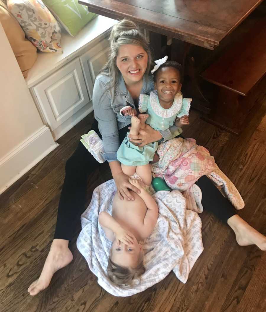 Mother sits on floor of kitchen with adopted African American daughter in her lap as she changes son's diaper