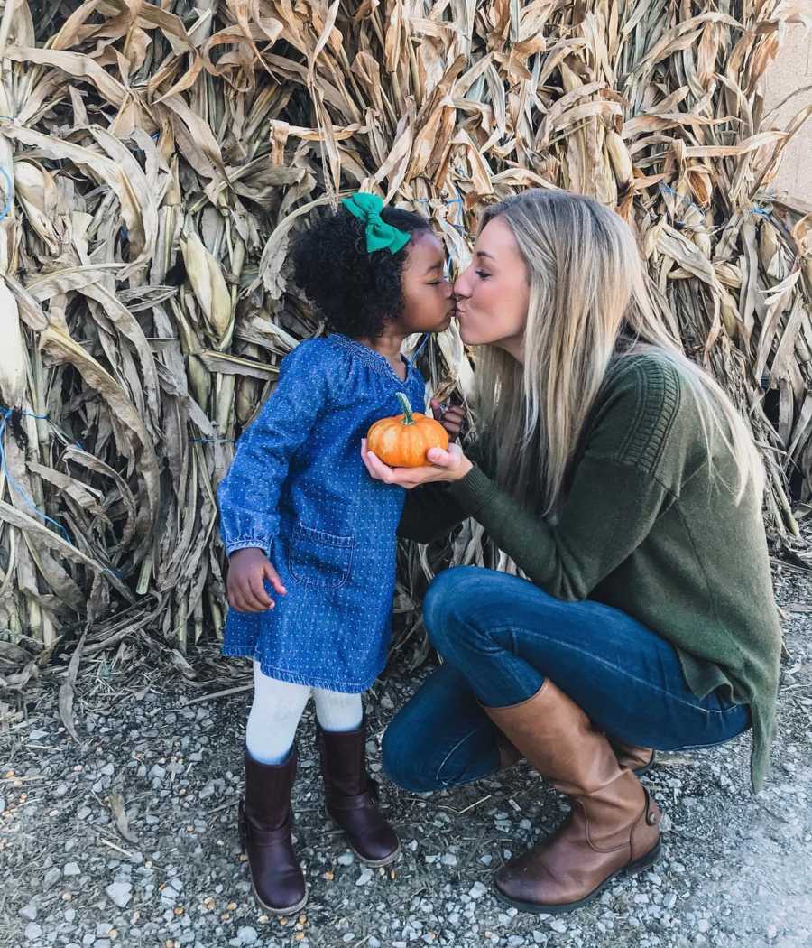 Mother kneels down to kiss adopted daughter while holding pumpkin in front of corn field