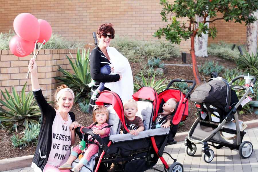 Mother squats down in front of stroller with triplets in it while grandmother stands in background holding fourth child