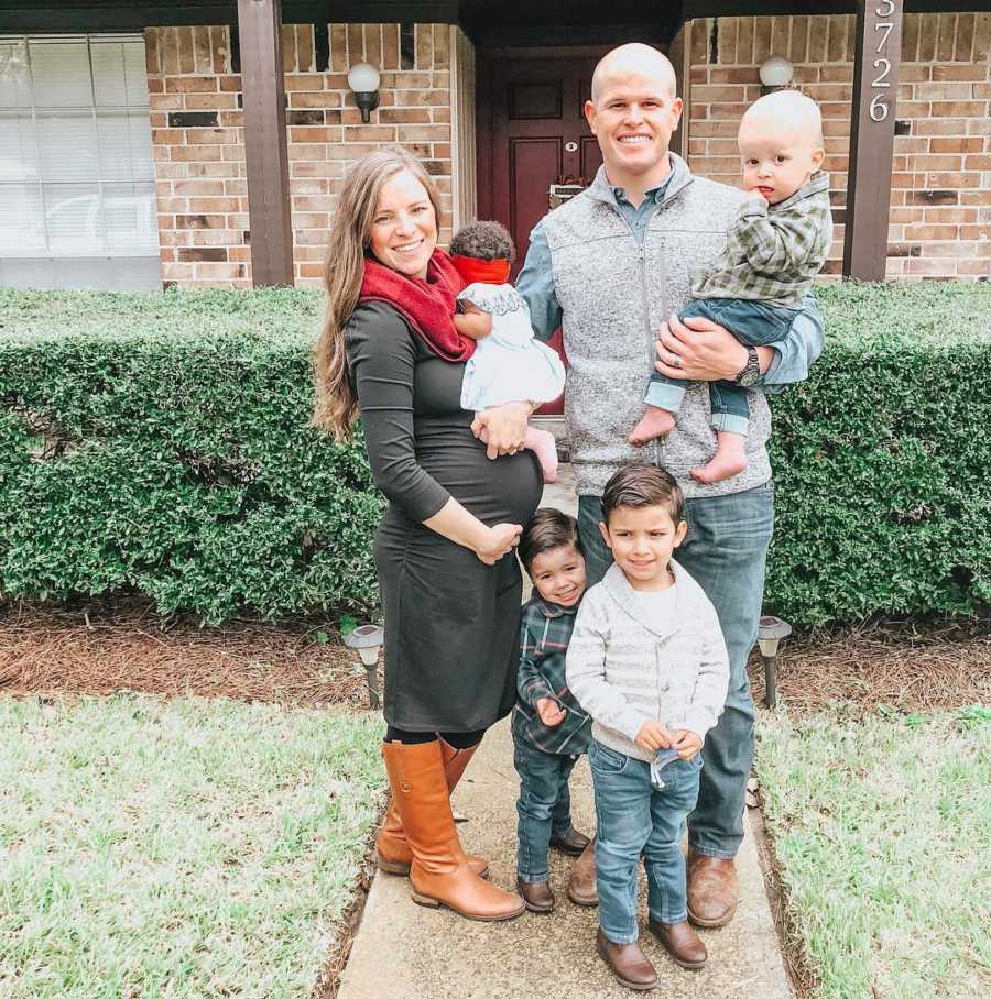 Pregnant wife stands outside of home holding foster baby beside husband and their three sons