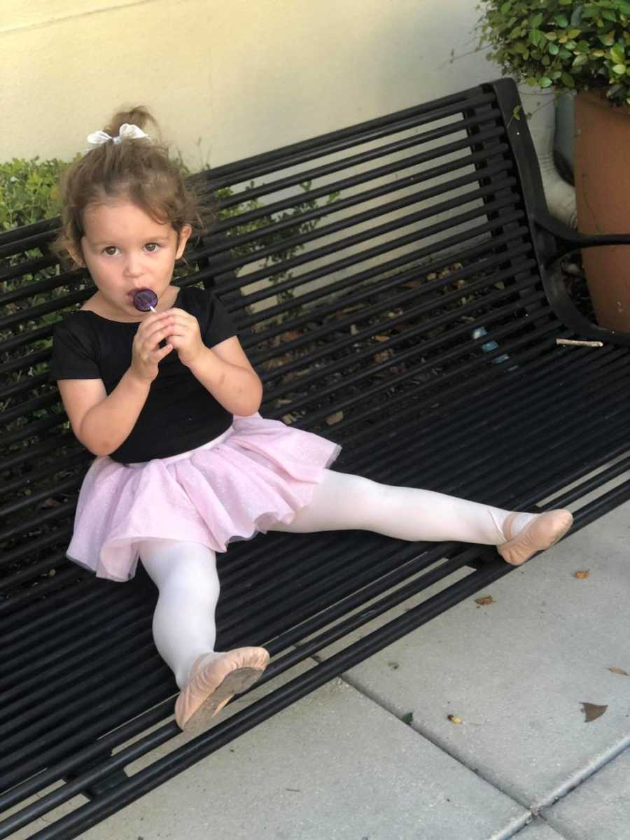 Little girl in ballet clothes sits on bench licking lollipop
