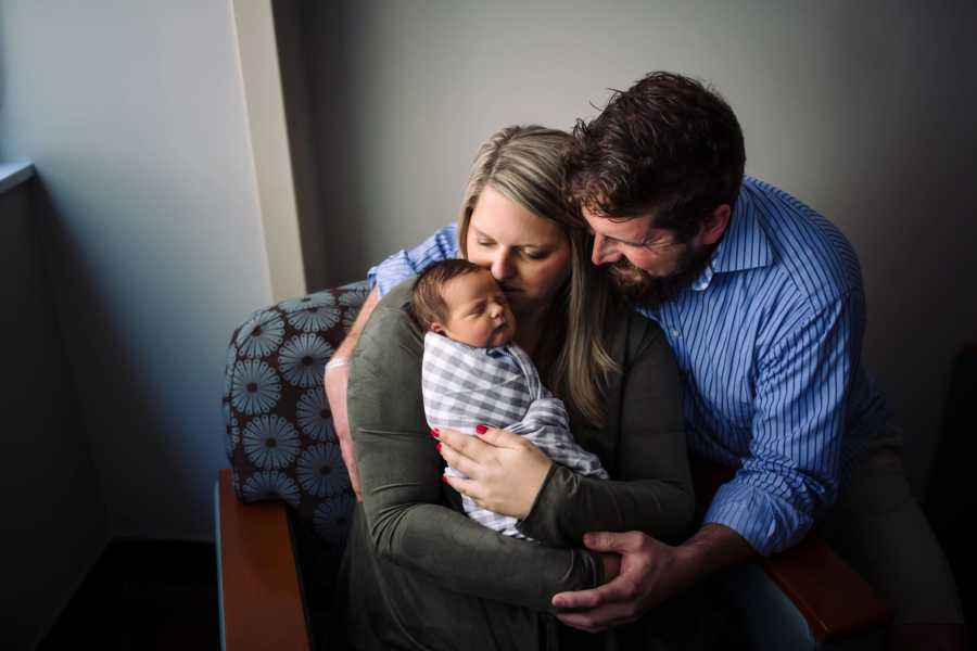 Mother sits in chair holding swaddled baby while husband leans over hugging wife