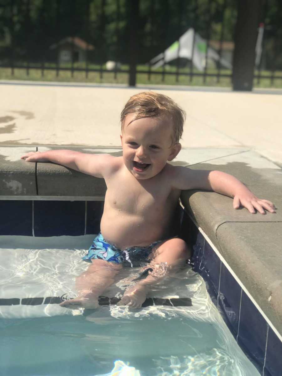 Toddler boy smiles as he sits on step of pool with arms out beside him resting on pool deck