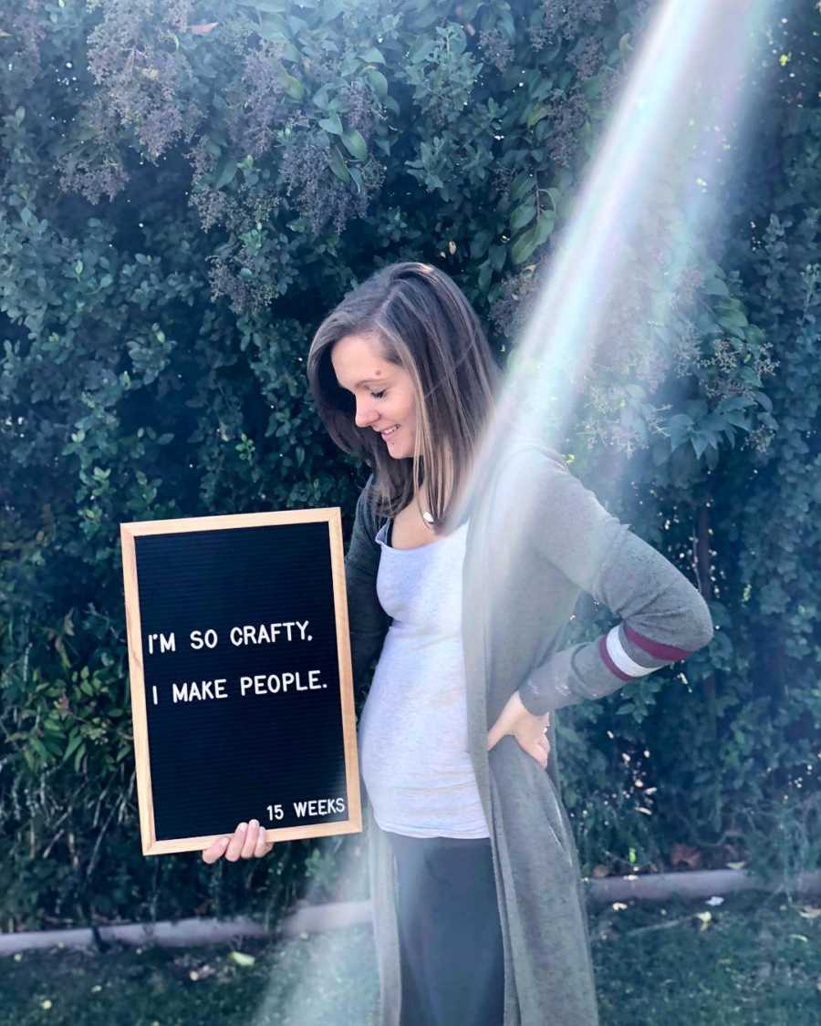 Pregnant woman stands outside smiling at sign she holds that says, "I'm so crafty I make people"