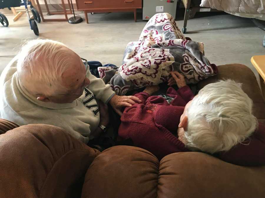 Elderly man sits on couch leaning over touching wife with dementia