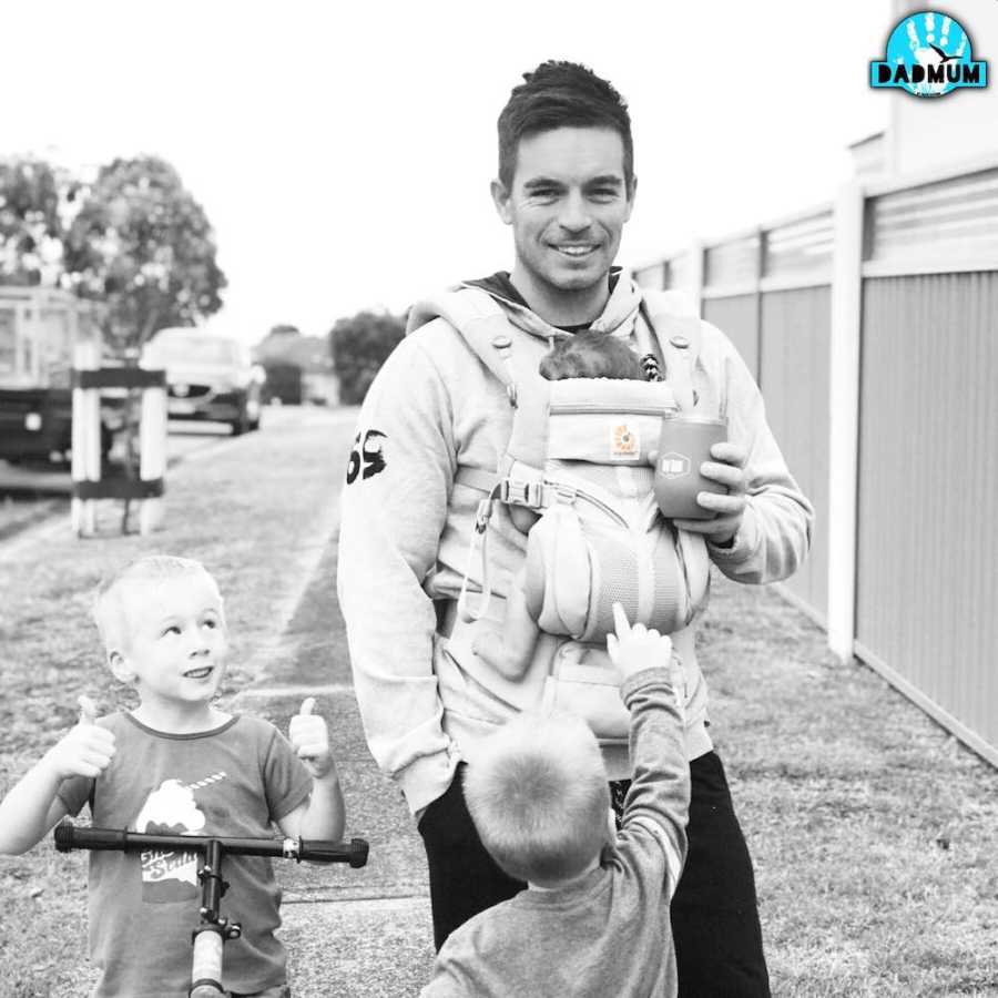 Man stands smiling outside with baby swaddled to his chest and two young boys by his side