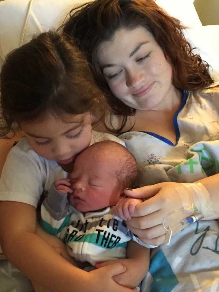 Mother who will have postpartum depression lays in hospital bed with firstborn who holds newborn in her lap