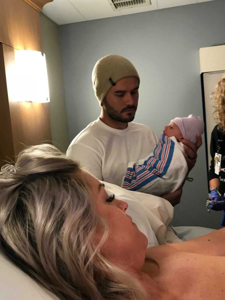 Husband stands in hospital room holding newborn beside wife who is laying in hospital bed