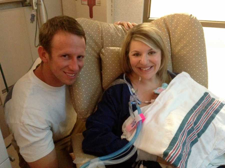 Mother sits in chair in NICU holding preemie baby under blanket while her husband kneels beside her