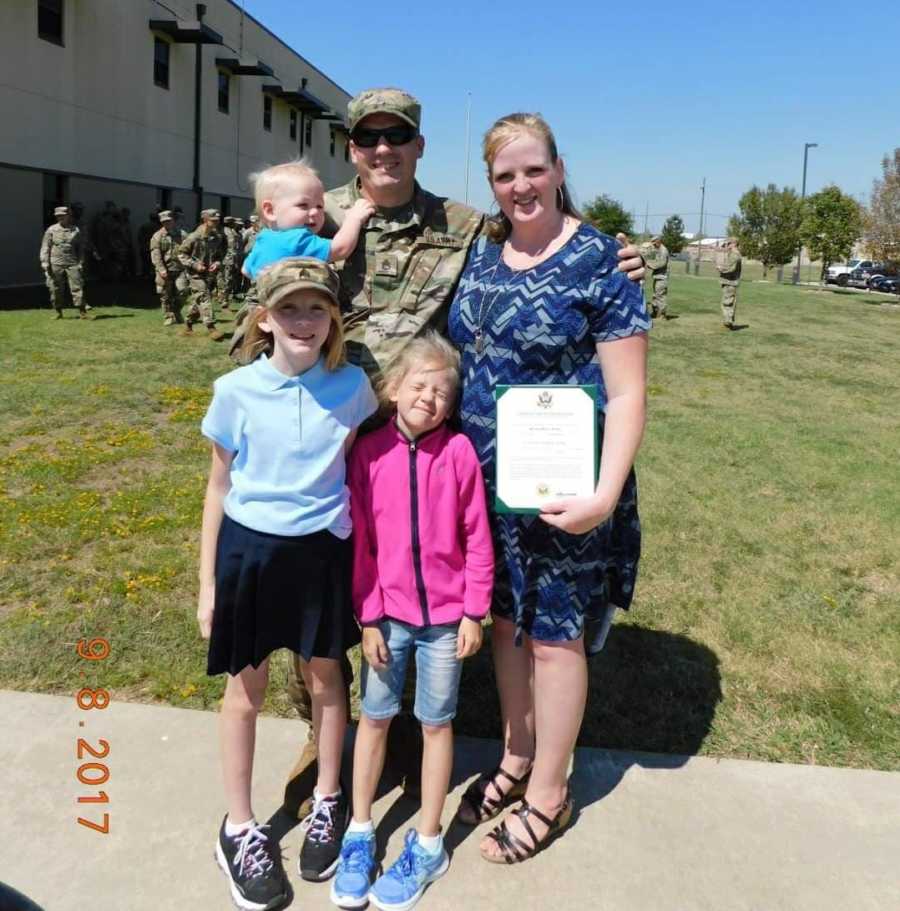 Active duty service member stands holding baby beside wife and two young daughters outside