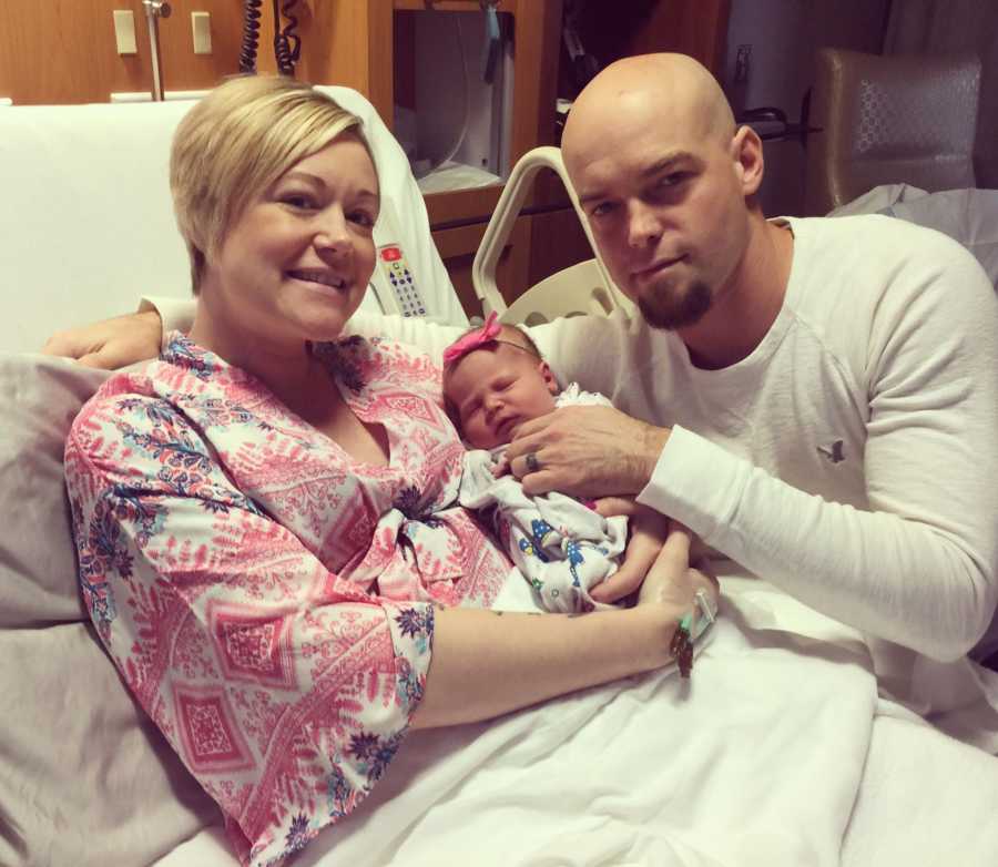 Woman lays in hospital bed with newborn who was conceived from egg donor while husband kneels beside them