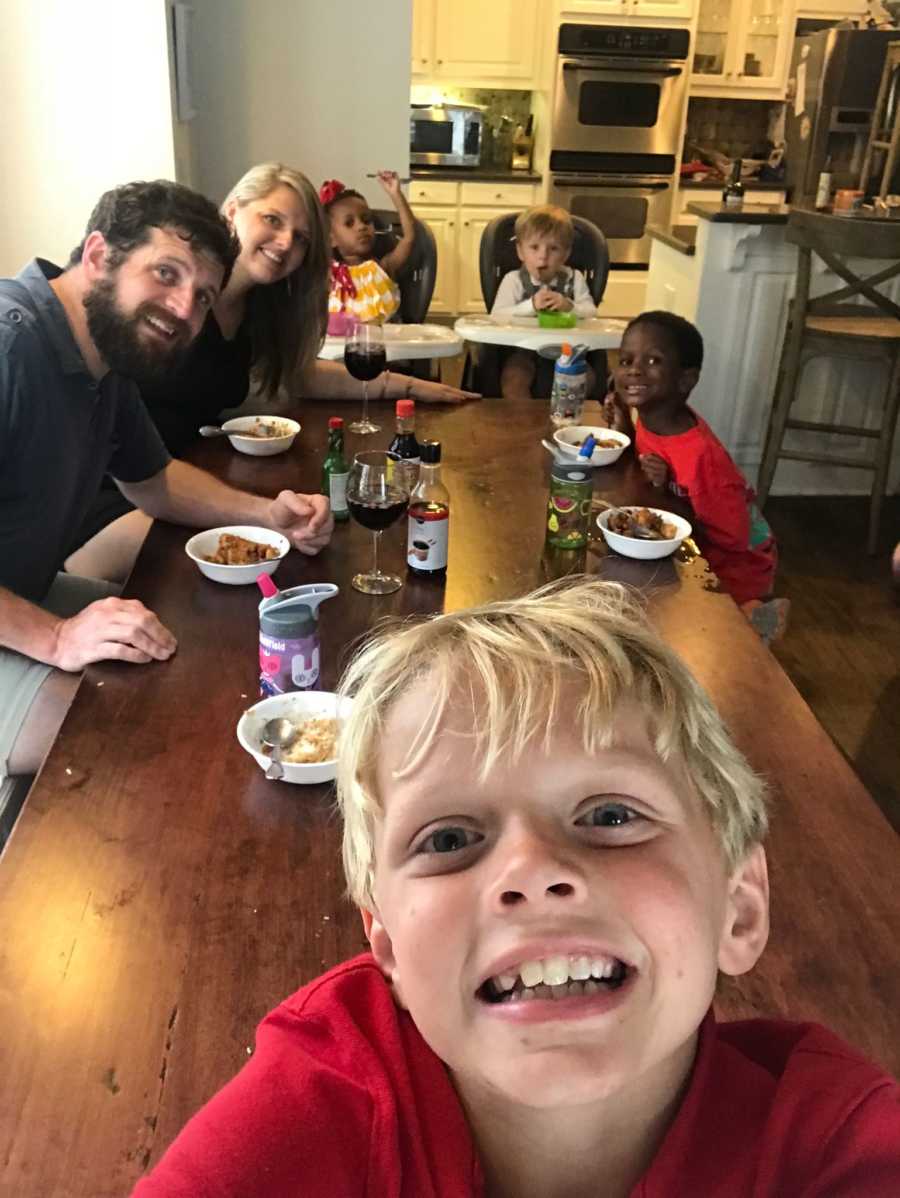 Young boy smiles in selfie with his mother, father, brother, and two adopted sisters sitting at dinner table 