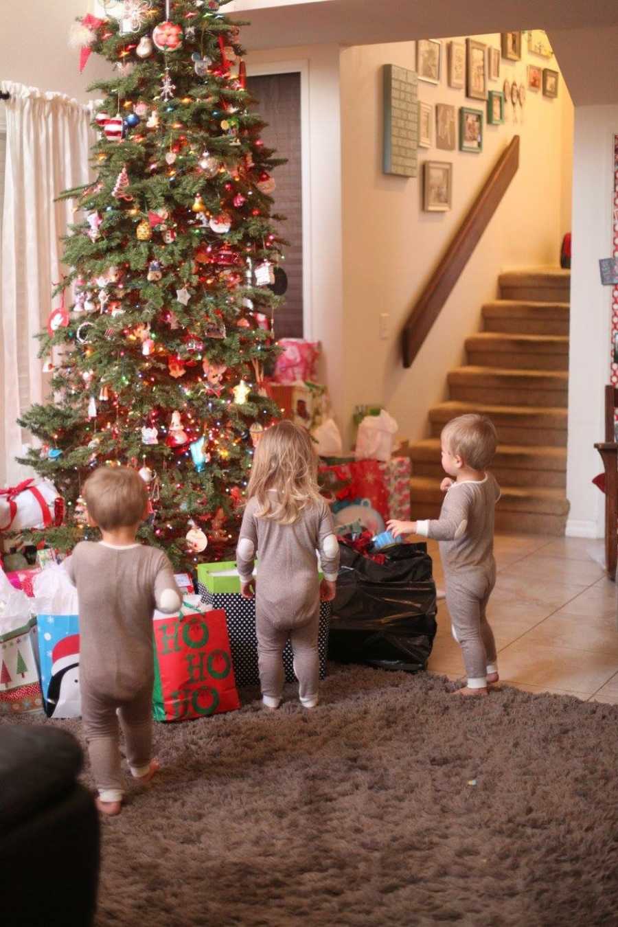 Triplets standing in matching onesies beside Christmas presents and tree in home