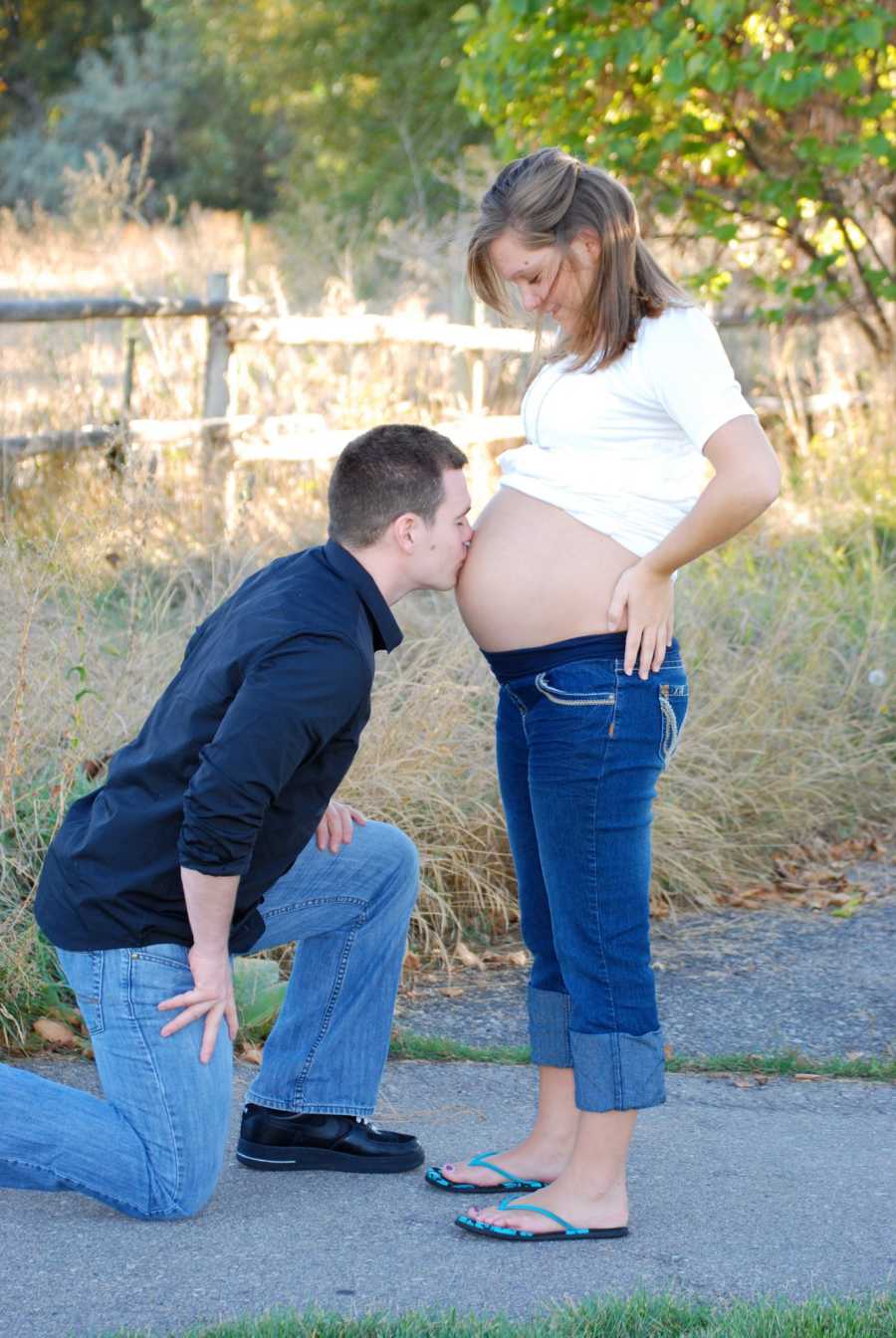 Pregnant woman stands outside with hands on her hips while husband kisses her stomach