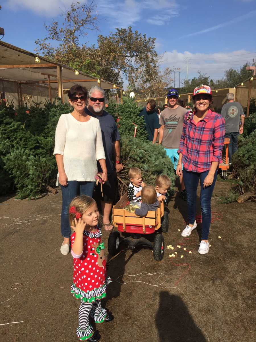 Husband and wife stand beside red wagon where their triplet grandchildren sit beside fourth grandchild 