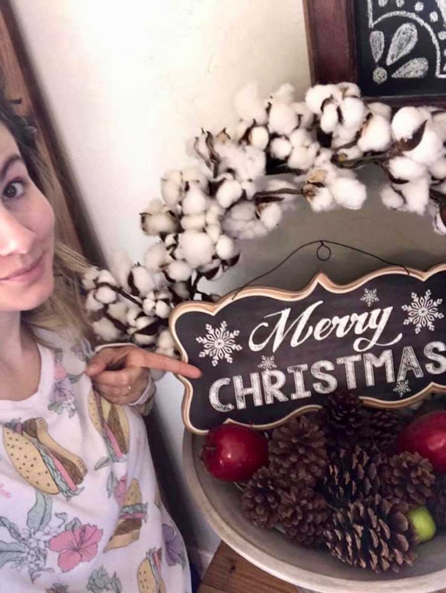 Mother smiles in selfie as she points to sign that says, "Merry Christmas"