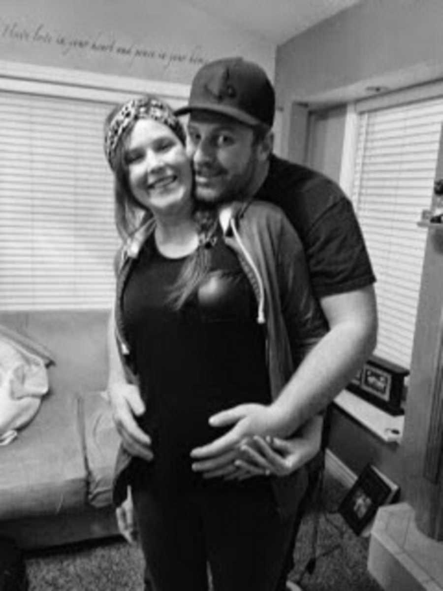 Husband stands smiling with arms wrapped around pregnant wife who ended up losing the baby