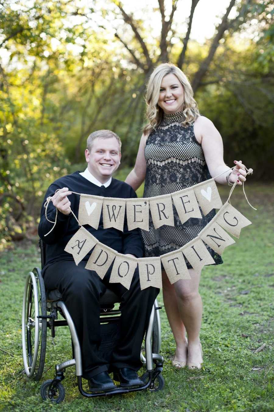 Husband in wheelchair holds sign with wife who stands beside him saying, "We're adopting"