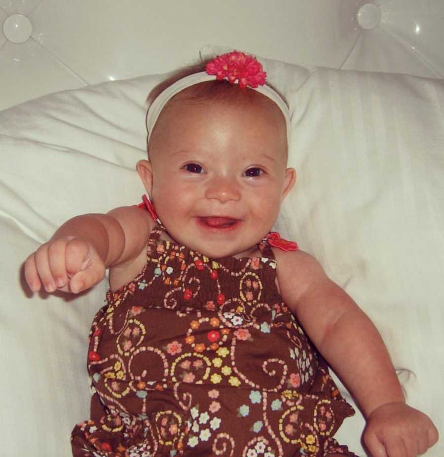 Baby with down syndrome lays smiling on her back in brown dress and headband with flower 